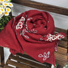 Ein tiefrotes, floral besticktes Wintertuch ist gerollt und auf einer Holzoberfläche drapiert.