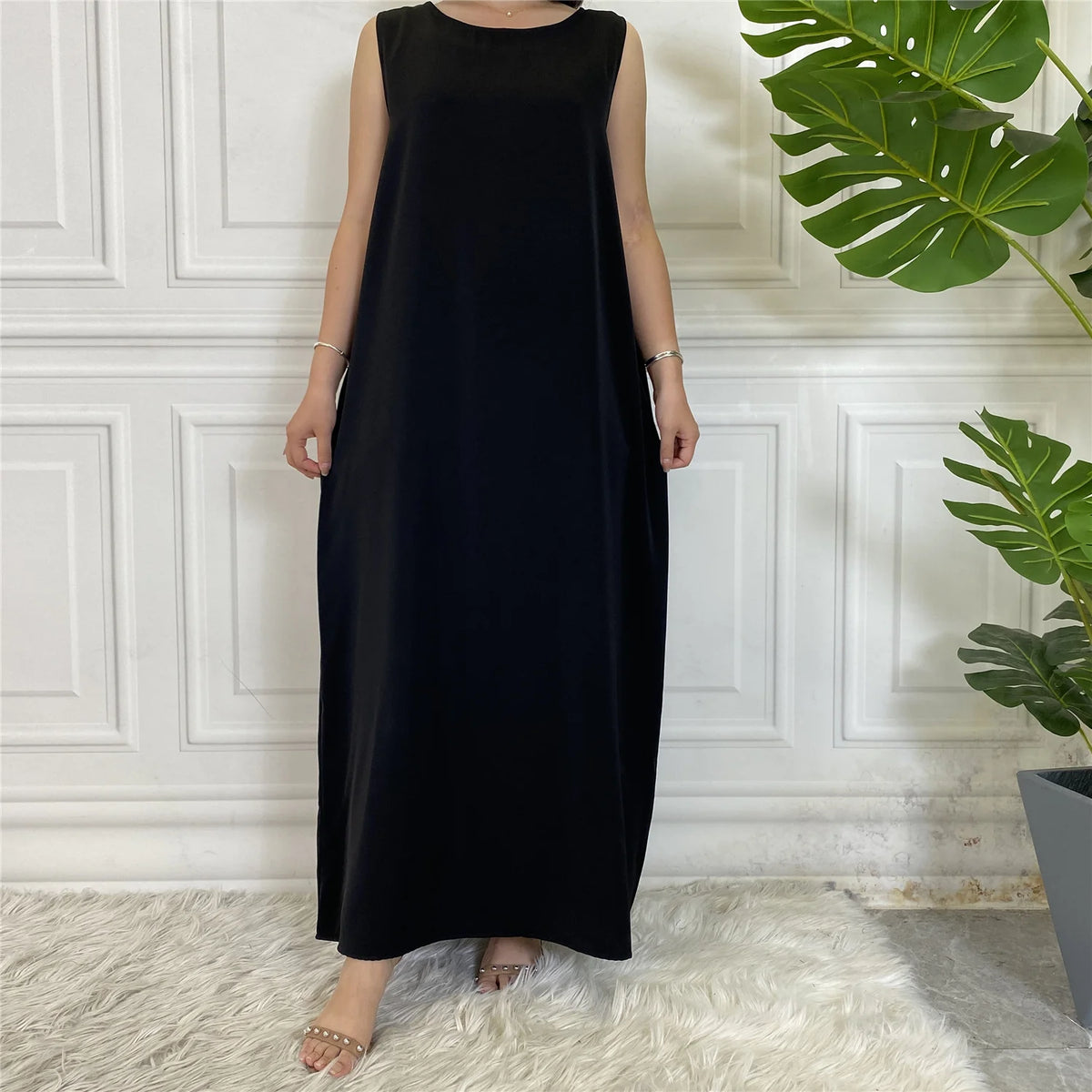 Langes schwarzes ärmelloses Kaftankleid auf einer Person vor weißer Wand mit großen grünen Blättern