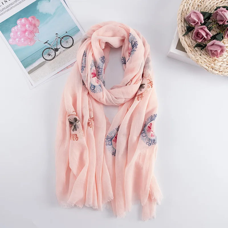 Weiches, pfirsichfarbenes, besticktes Wintertuch mit Blumenmuster, drapiert auf weißer Fläche neben einer Postkarte.