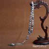 A dark wooden stand holds a string of grey and white agate prayer beads with a silver tassel necklace.