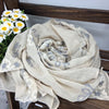 Beiger, bestickter Winter-Schal mit Blumen, gerollt und auf Holzoberfläche mit weißen Blumen drapiert.