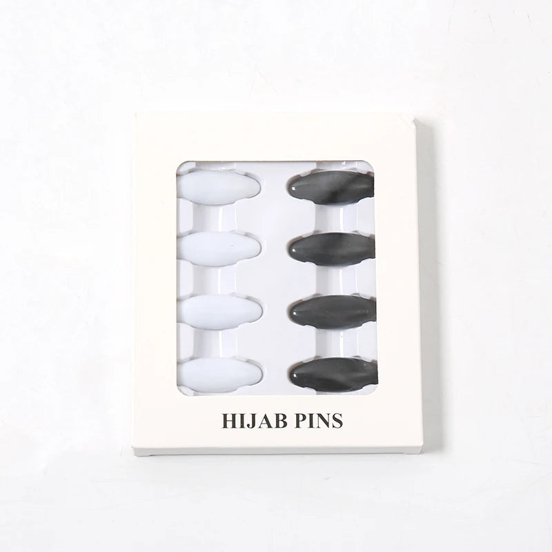 Una caja rectangular blanca que contiene ocho sujetadores y pinzas para pañuelos (hijabs) en blanco y negro.