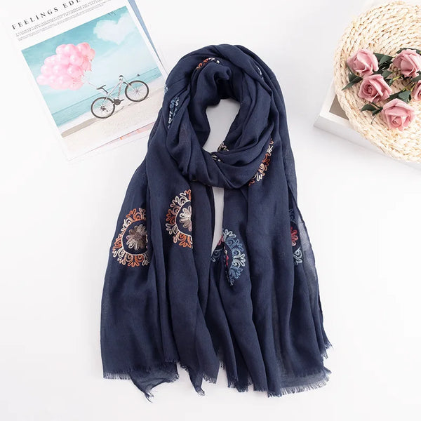 A dark blue floral embroidered winter scarf draped on a white surface next to a woven basket and a magazine.