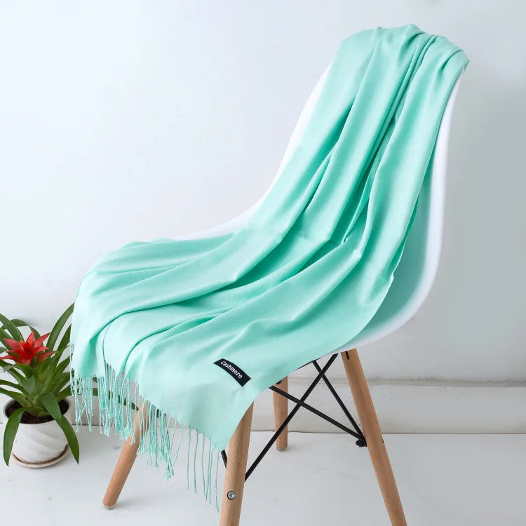 Un chal de cachemira verde menta colgado sobre una silla de madera, con detalles de flecos y una planta de fondo.