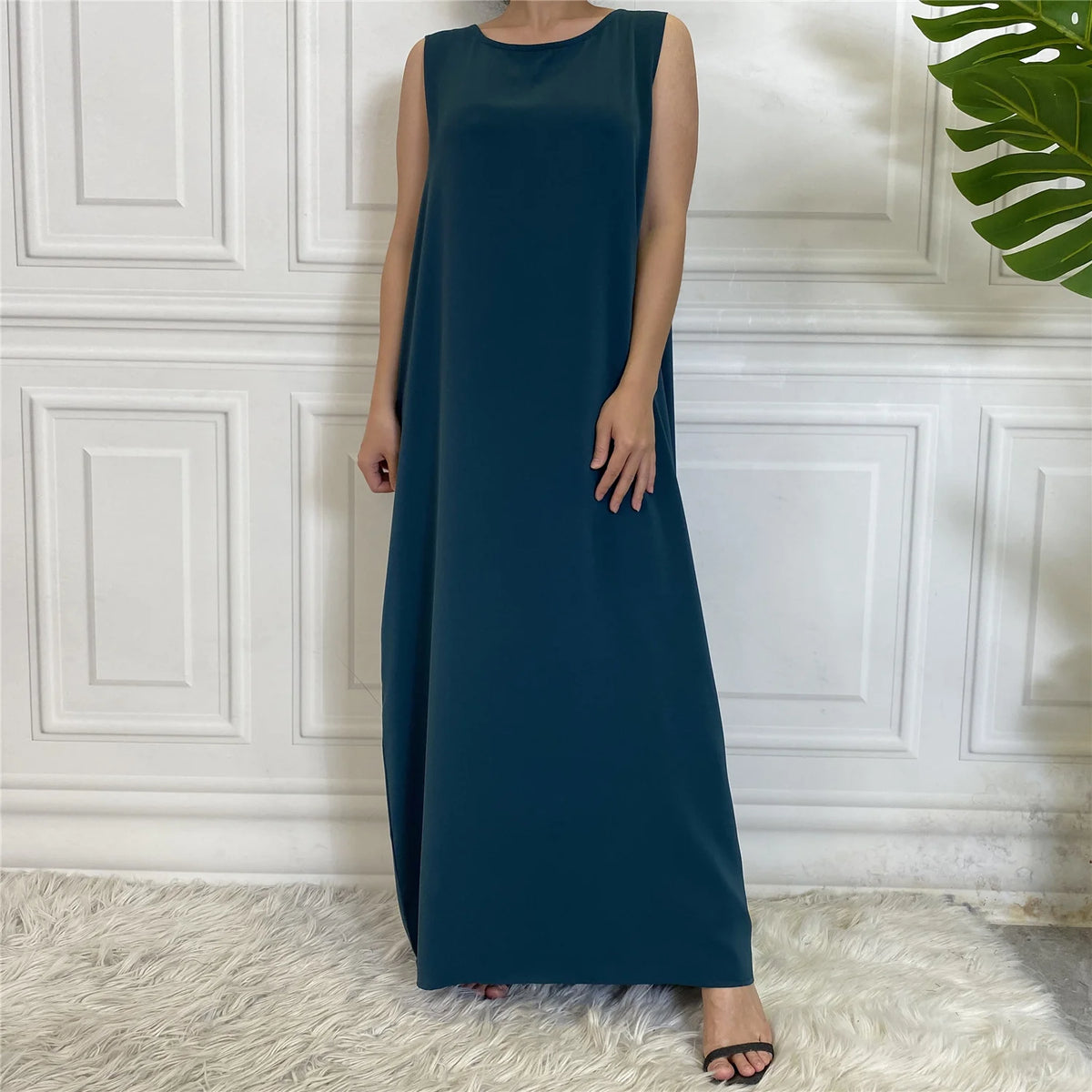 Dunkel-türkises ärmelloses, bedecktes Kaftankleid vor einer weißen Paneelwand.