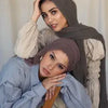 Eine Frau mit olivgrüner Chiffon-Hijab und heller Kleidung, die in die Kamera blickt.