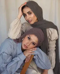 Eine Frau mit olivgrüner Chiffon-Hijab und heller Kleidung, die in die Kamera blickt.