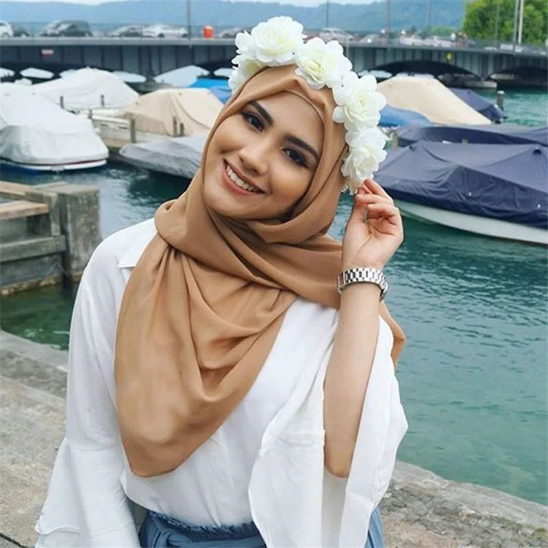 Eine Frau in beigefarbigem Chiffon-Hijab und weißem Oberteil posiert draußen in der Nähe von Wasser mit Booten im Hintergrund