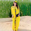 Eine Frau in einem leuchtend gelben Kaftan-Set mit farbenfroher Stickerei und einer gelben Blume im Haar, draußen stehend.