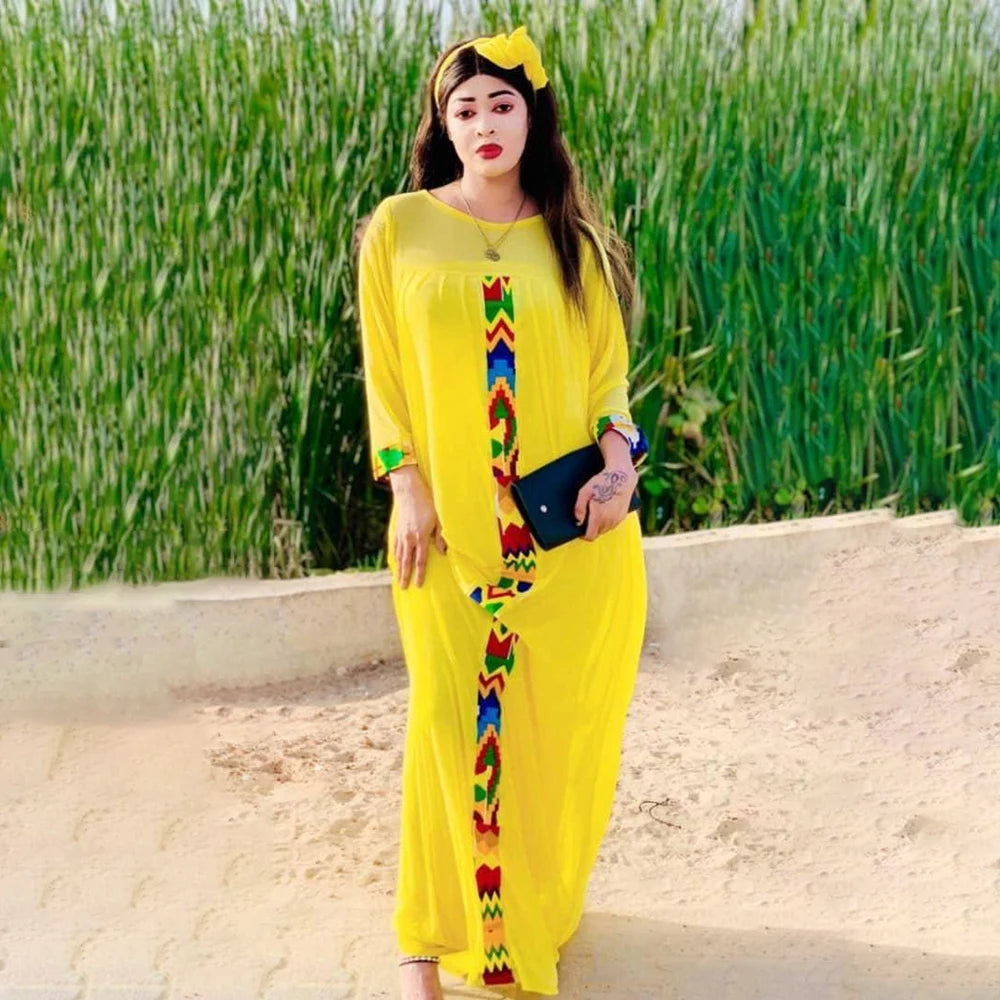 Eine Frau in einem leuchtend gelben Kaftan-Set mit farbenfroher Stickerei und einer gelben Blume im Haar, draußen stehend.
