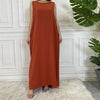 Eine Frau in einem orange-braunen ärmellosen, bescheidenen Kaftankleid vor einer weißen Paneelwand.