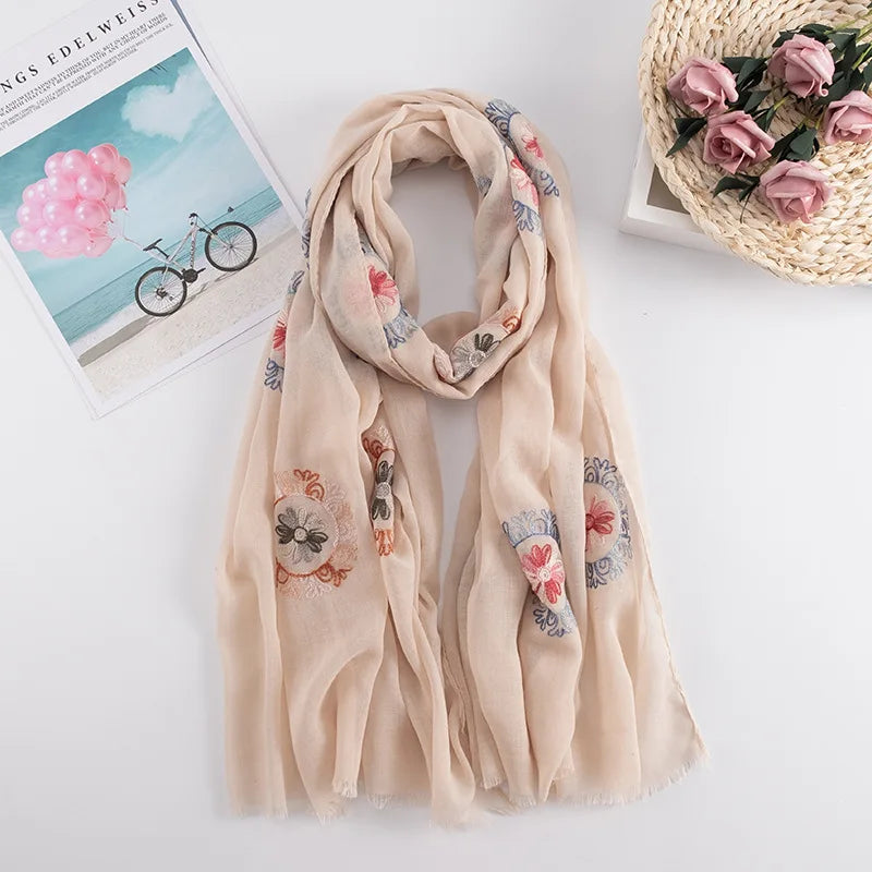 Beiger, bestickter Winter-Schal mit Blumenmuster auf weißem Hintergrund neben einer Postkarte.