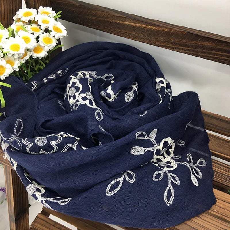 Dunkelblauer floraler bestickter Winterschal neben einem Strauß weißer und gelber Blumen.