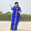 Eine Frau in einem königsblauen Kaftan-Set mit farbenfroher Stickerei steht draußen vor einer trockenen Landschaft.
