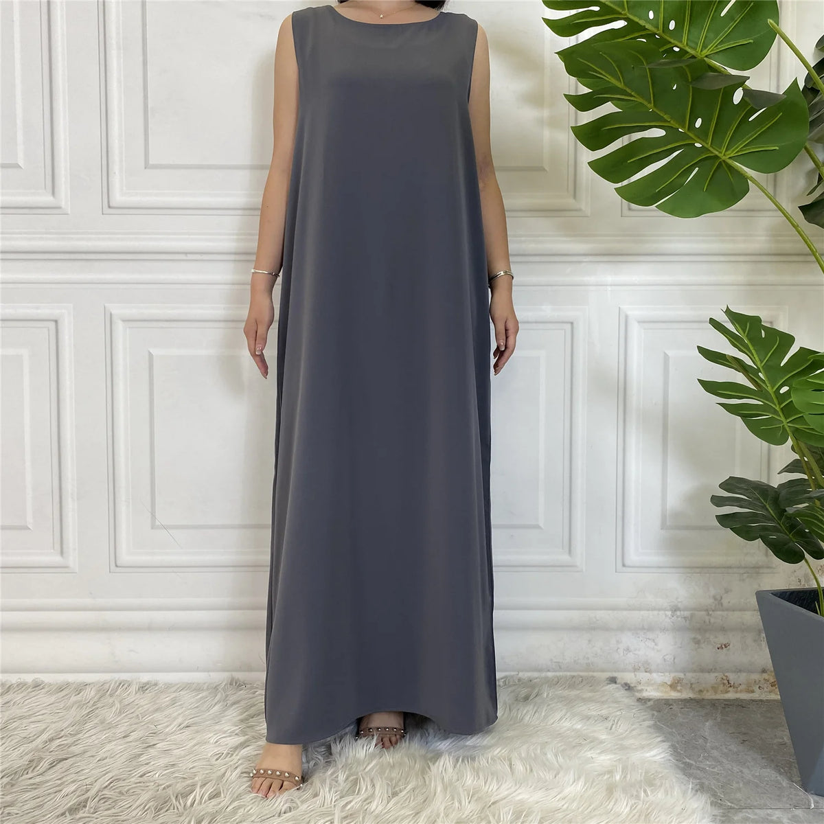 Langes, langärmelloses Kaftankleid in anthrazitgrauer Farbe vor einer weißen Paneelwand.