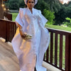 A woman wearing an elegant white kaftan with tatting detail standing outdoors by a wooden railing.