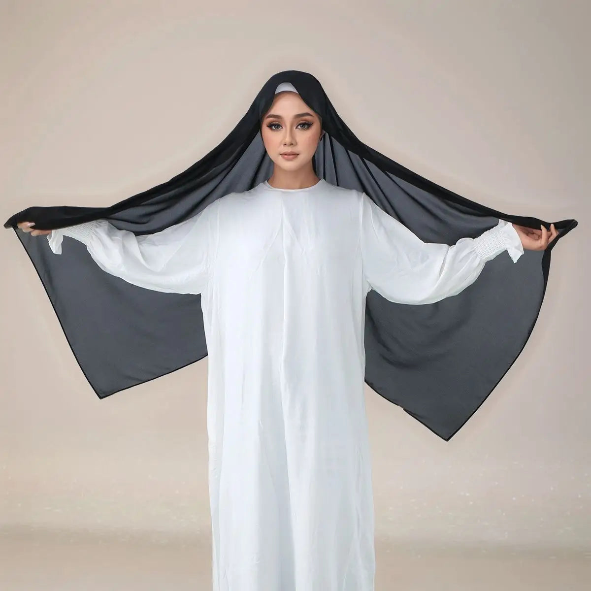 Eine Frau trägt ein weißes langes Kleid und einen schwarzen Instant-Hijab mit Riemchen, über ihren Kopf drapiert.