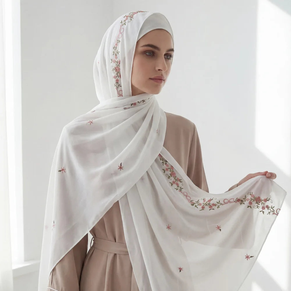 A woman wearing an elegant white chiffon hijab shawl with delicate floral embroidery, draped over a beige outfit.