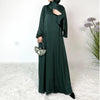 Una mujer vistiendo un vestido maxi de satén verde oscuro y hijab, de pie en interiores.