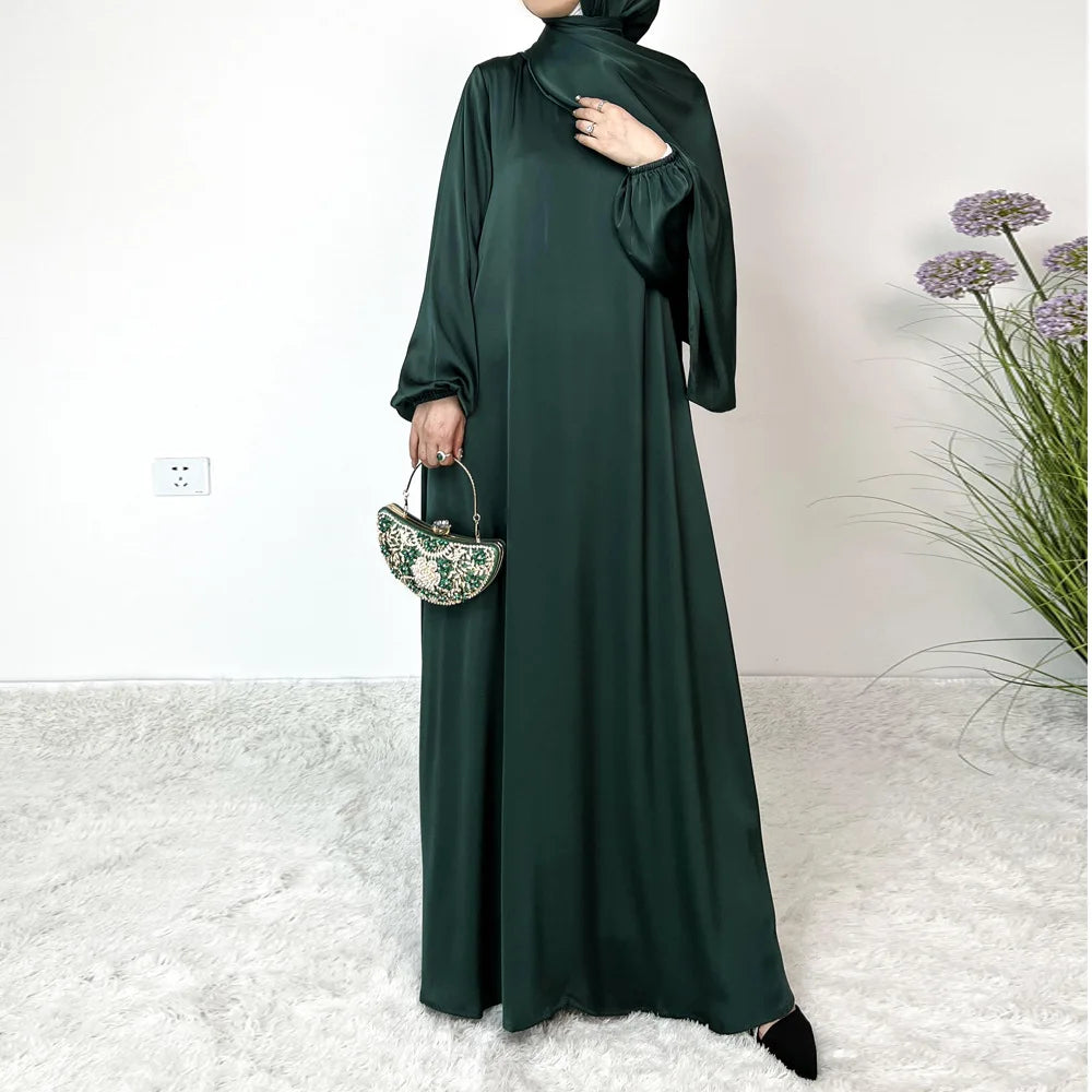 Una mujer vistiendo un vestido maxi de satén verde oscuro y hijab, de pie en interiores.