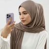 A woman wearing a beige hijab and white top is holding a purple smartphone.