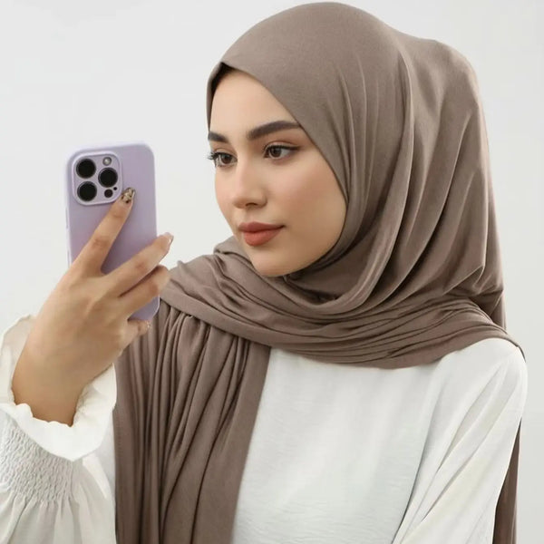 A woman wearing a beige hijab and white top is holding a purple smartphone.