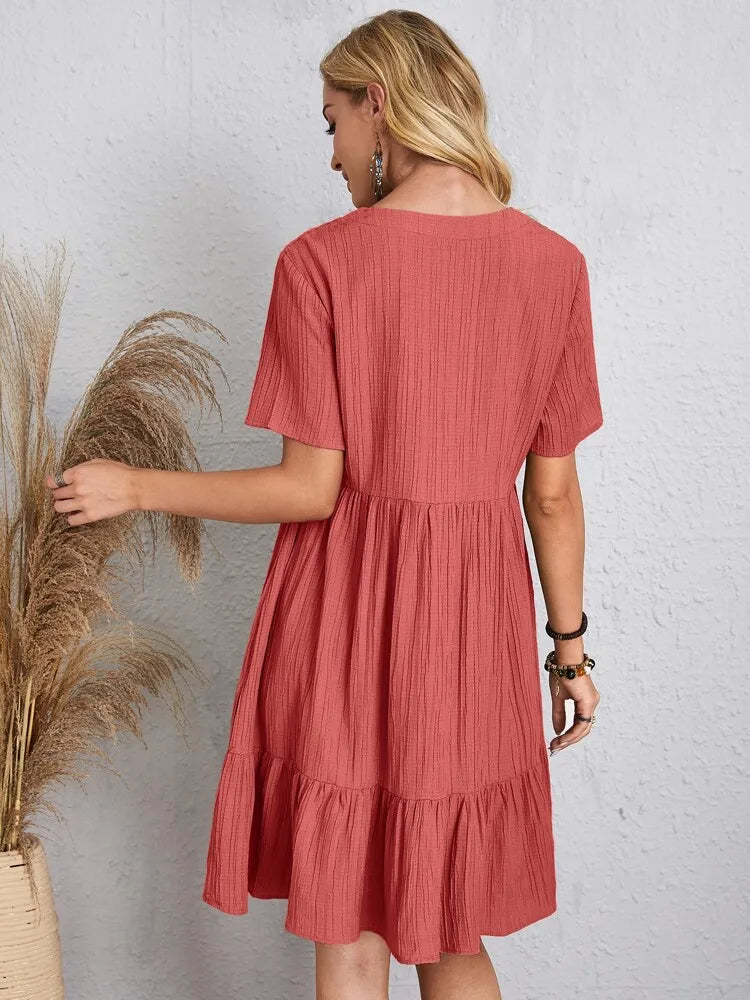 Eine Frau trägt ein korallenrosa, kurzärmeliges A-Linien-Minikleid mit vertikaler Rippung vor einer hellgrauen Wand.