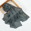 A gray chiffon hijab shawl with delicate pink floral embroidery, rolled and draped on a wooden frame.