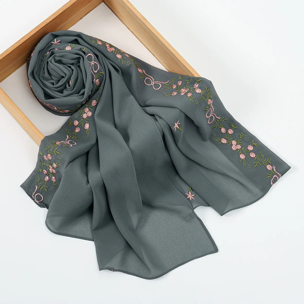 A gray chiffon hijab shawl with delicate pink floral embroidery, rolled and draped on a wooden frame.
