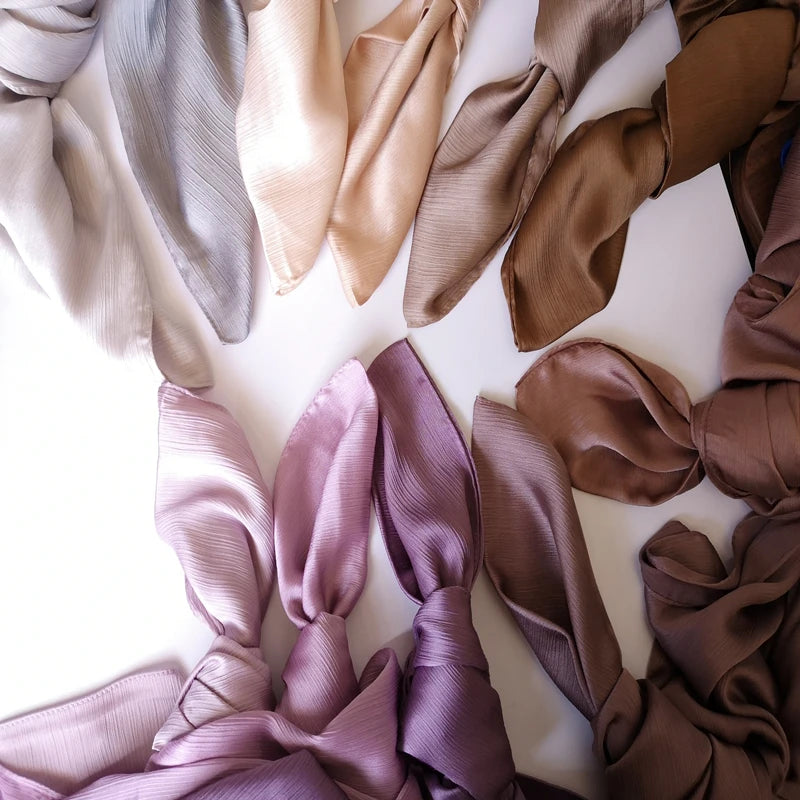 Eine Sammlung von Crinkle-Satin-Chiffon-Hijabs in verschiedenen neutralen und mauvenfarbenen Tönen auf einer weißen Oberfläch