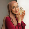 A woman wearing a beige chiffon hijab scarf and a maroon top, holding a glass perfume bottle with a floral stopper.