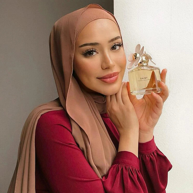 A woman wearing a beige chiffon hijab scarf and a maroon top, holding a glass perfume bottle with a floral stopper.
