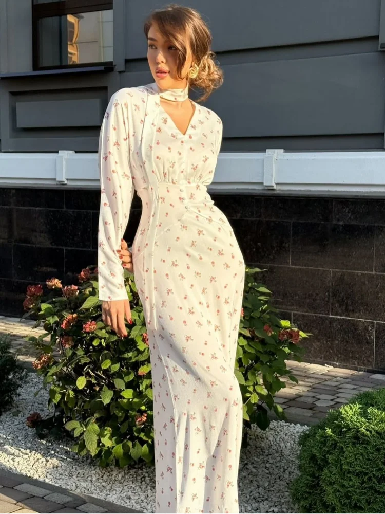 Una mujer vistiendo un vestido largo de satén con estampado floral blanco, de pie al aire libre contra una pared gris.