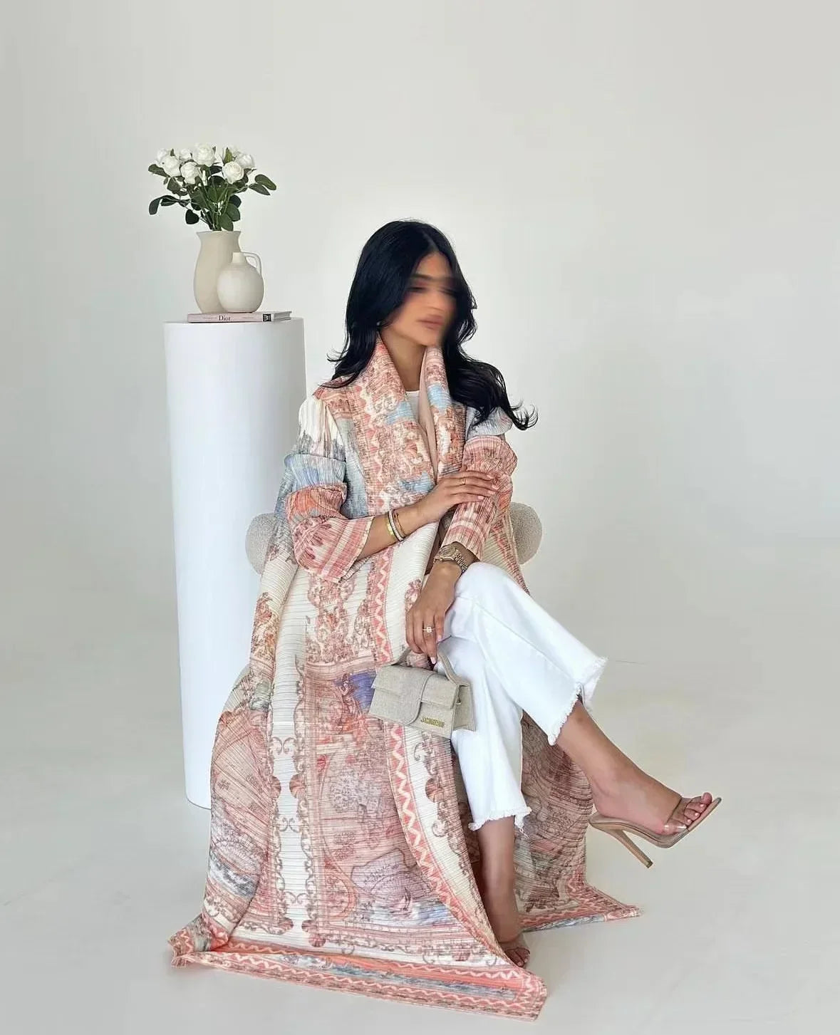 Femme portant un trench long de couleur rose et blanc à plis avec un pantalon blanc, posant à côté d'un vase de fleurs blanch