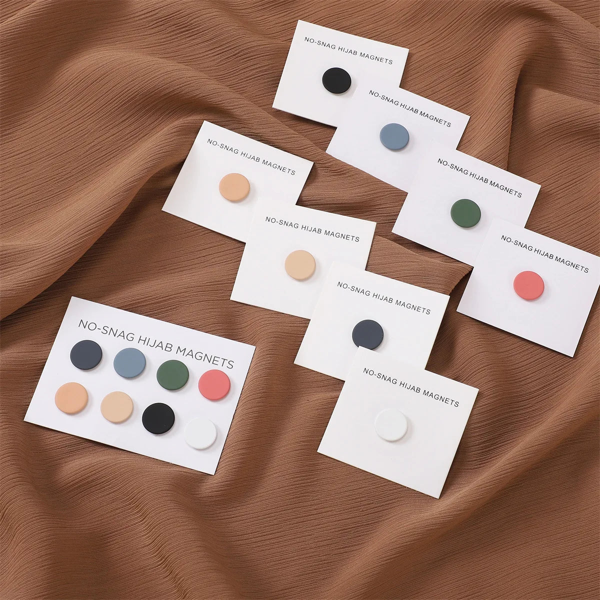 Colección de pines/broches magnéticos de hijab coloridos en tonos rosa, verde y gris mostrados sobre tela marrón