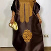 Eine Frau in einem dunkelbraunen traditionellen Ankara Dashiki-Kleid mit Goldstickerei und Schmuck.