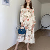 Eine Frau in einem weißen floralen Maxi-Kleid und einer blauen Handtasche in einem modernen Raum.