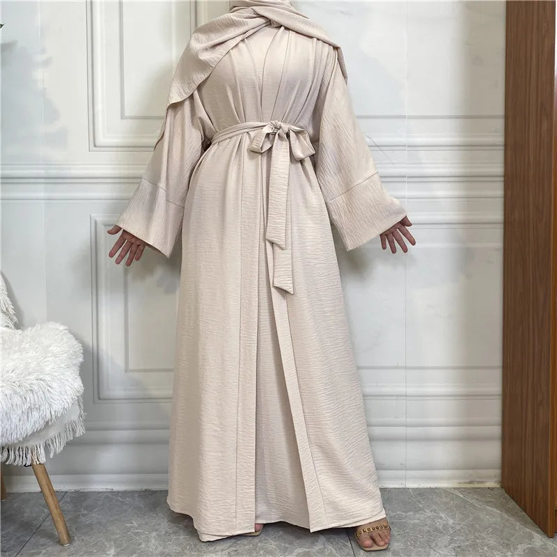 A beige elegant women's abaya and kaftan set worn by a model standing in front of a white paneled wall.