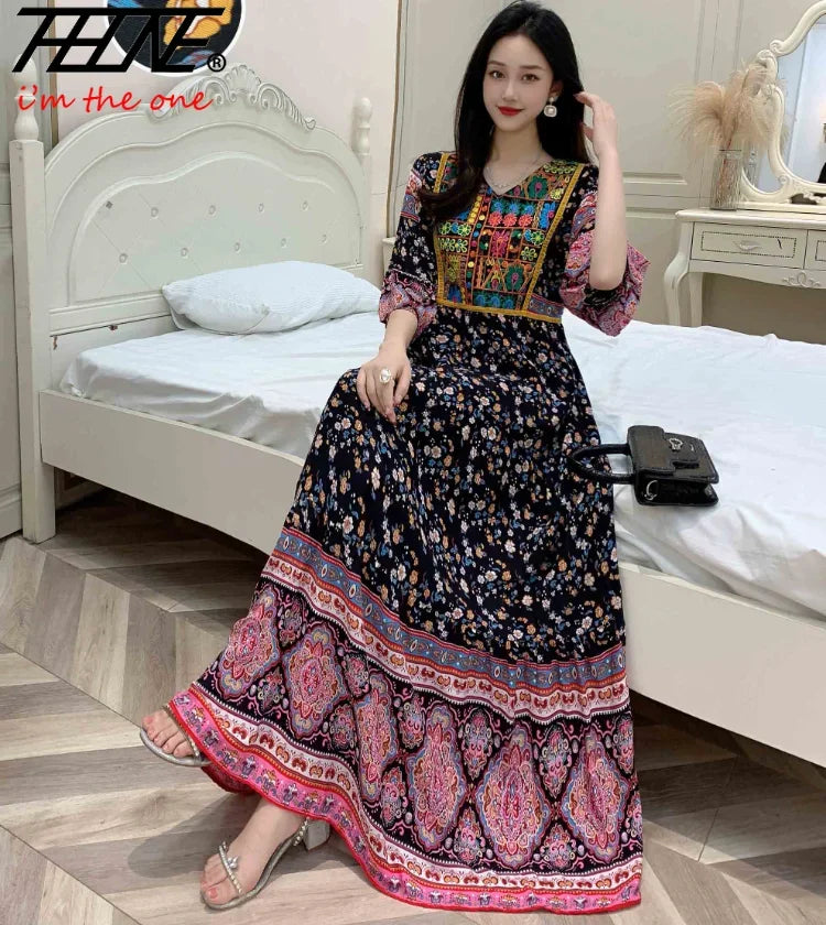 Eine Frau modelliert ein langes, dunkles, besticktes Maxi-Kleid mit farbigem Zierrand in einem Schlafzimmer.