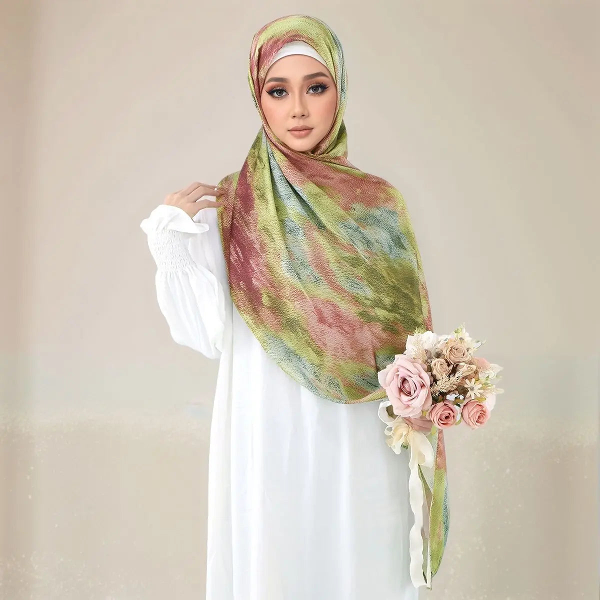 Eine Frau trägt einen elegant gemusterten Seideschal-Hijab in Grün-, Pink- und Brauntönen über einem weißen Kleid.