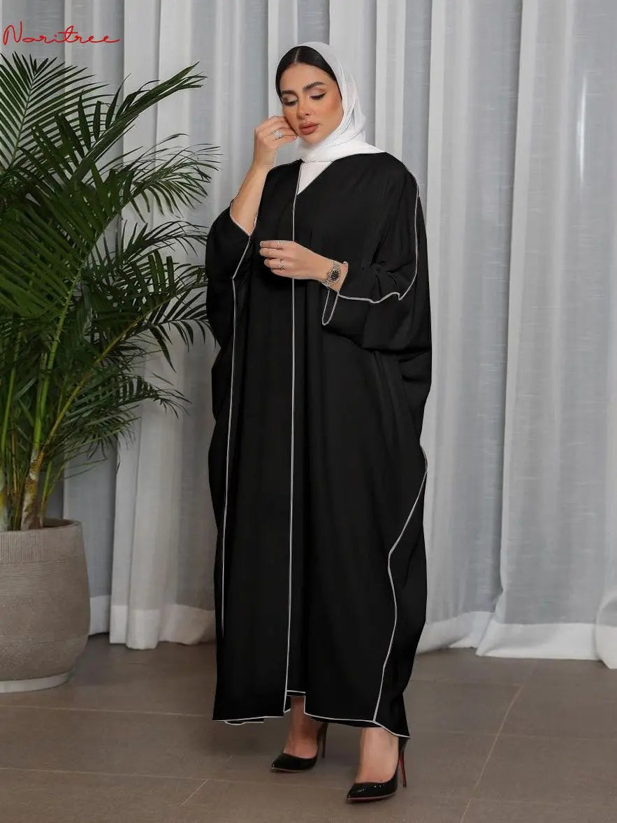 Una mujer con un abaya negro oversize y ribetes blancos, y un hijab blanco, de pie frente a una cortina transparente.