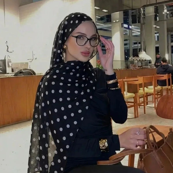 A woman wearing a black polka dot shawl and glasses sits in a modern indoor setting.