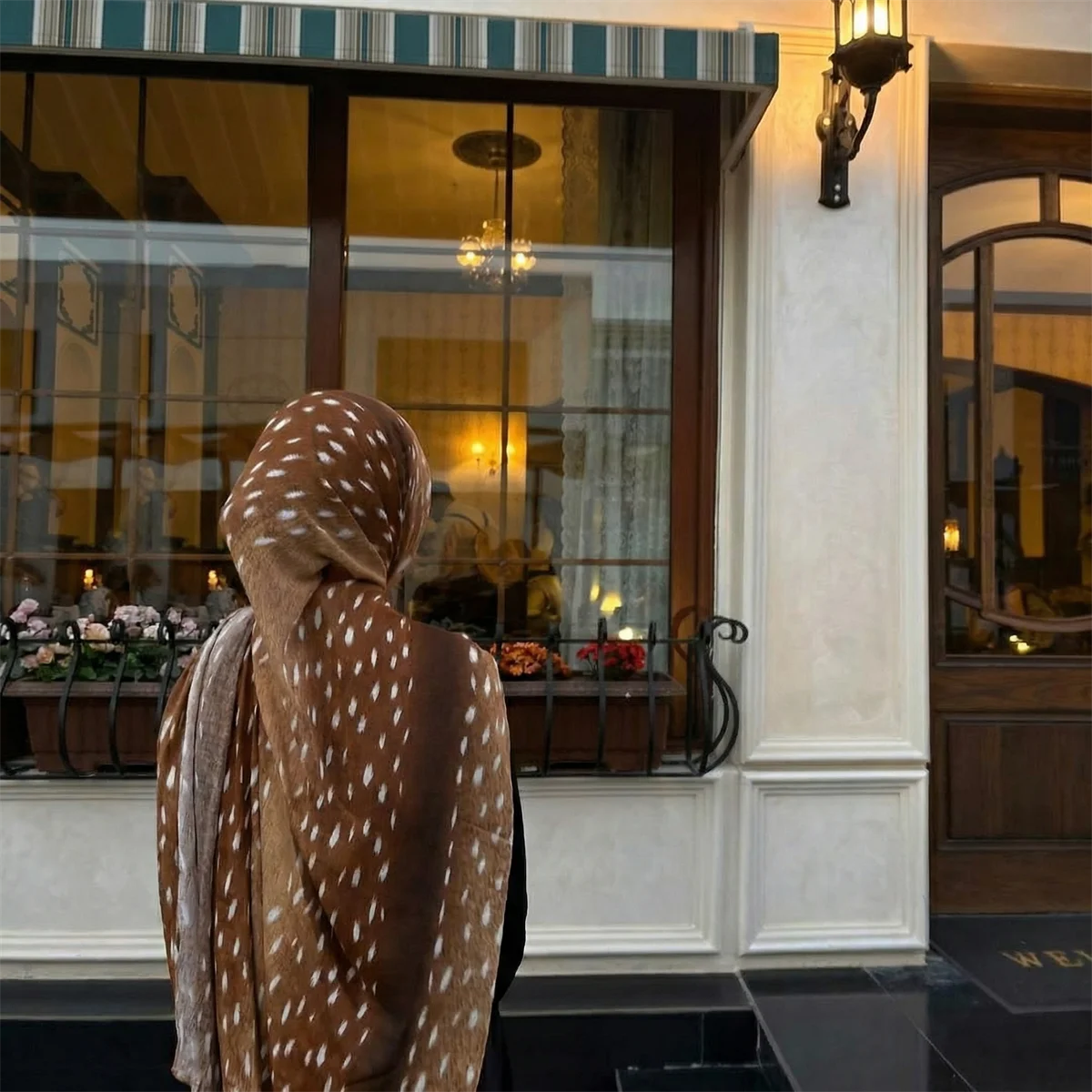 Eine Frau mit braunem Hirschmuster-Hijab-Schal steht vor einem Glasfenster.