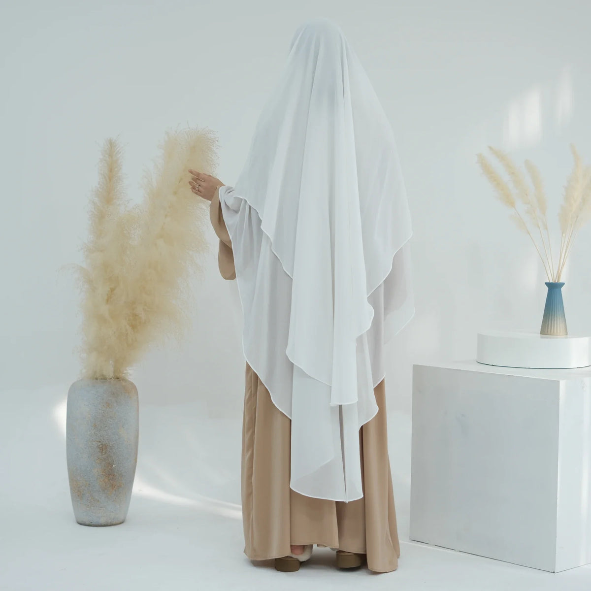 Eine Frau trägt einen weißen, zweilagigen Chiffon-Niqab-Kopftuch neben getrocknetem Ziergras in einer Vase.