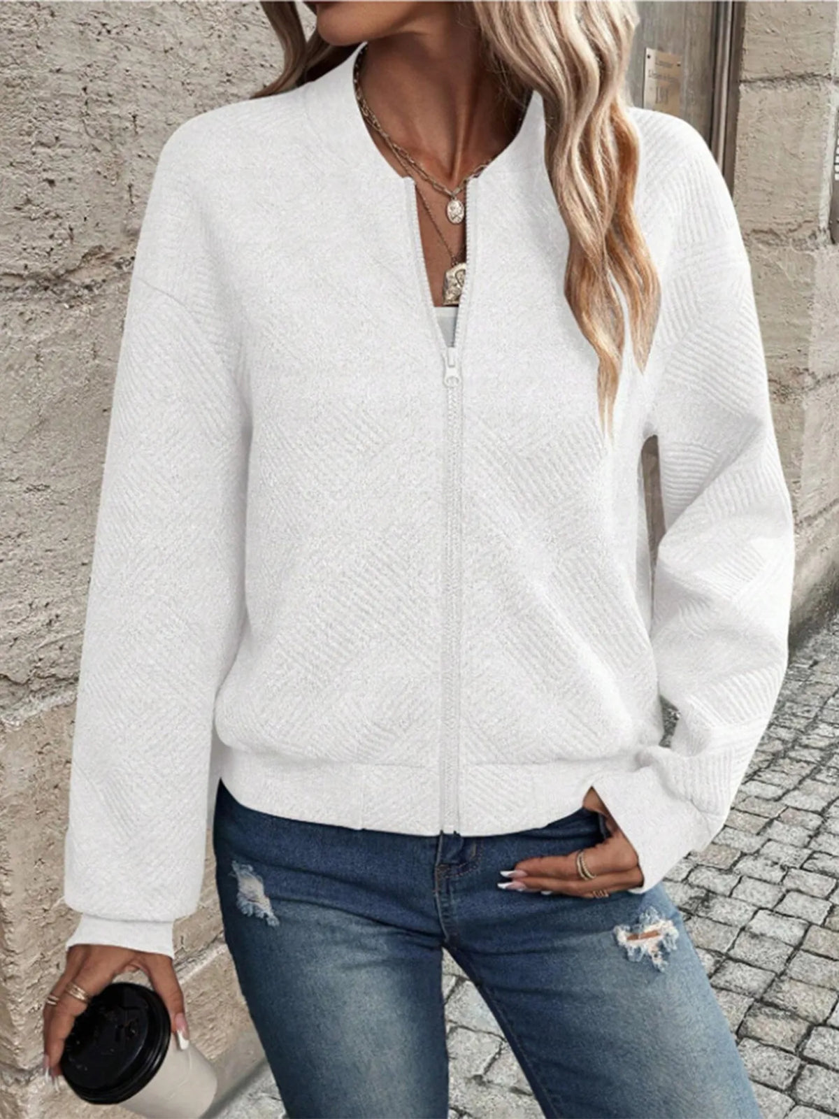 Une femme portant une veste zippée en tricot blanc et un jean bleu, debout à l'extérieur.