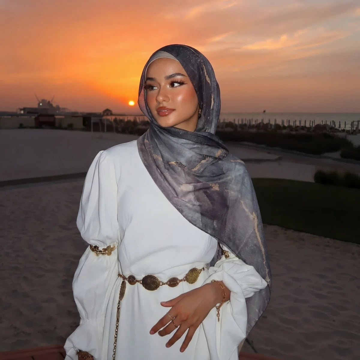 Eine Frau in weißer Kleidung und grauem Aquarell-Hijab vor einem Sonnenuntergang.