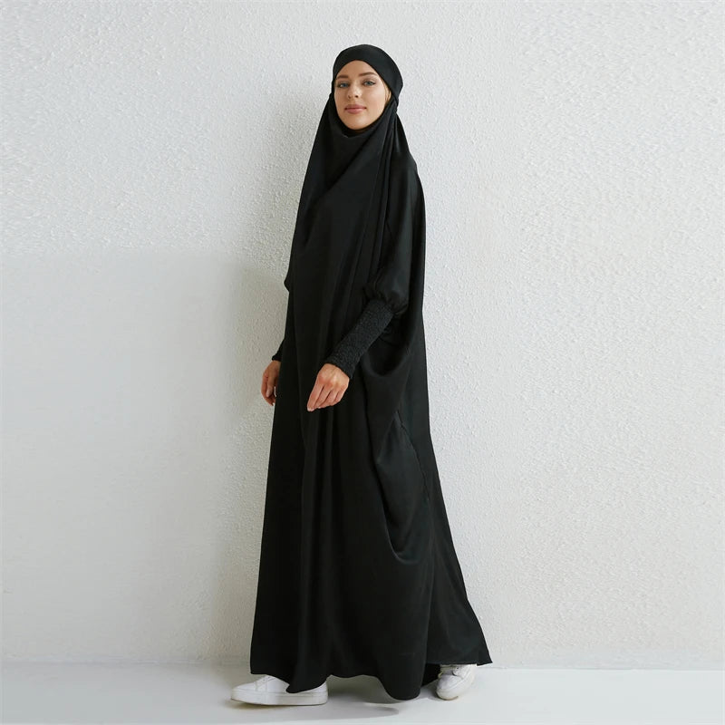 Eine Frau in einem schwarzen, bescheidenen Abaya mit gerafften Ärmeln vor einer weißen Wand.