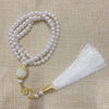 Weiße Perlenrosary-Armband mit goldener Anhänger und weißem Quaste auf beigem Stoff.