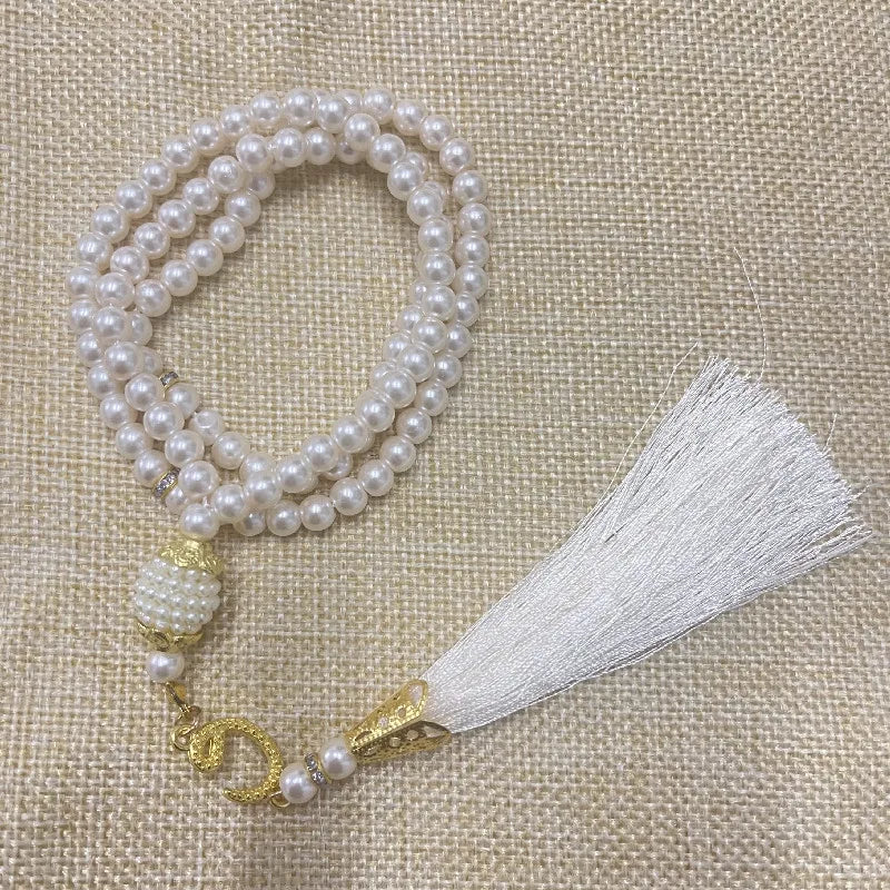 Weiße Perlenrosary-Armband mit goldener Anhänger und weißem Quaste auf beigem Stoff.