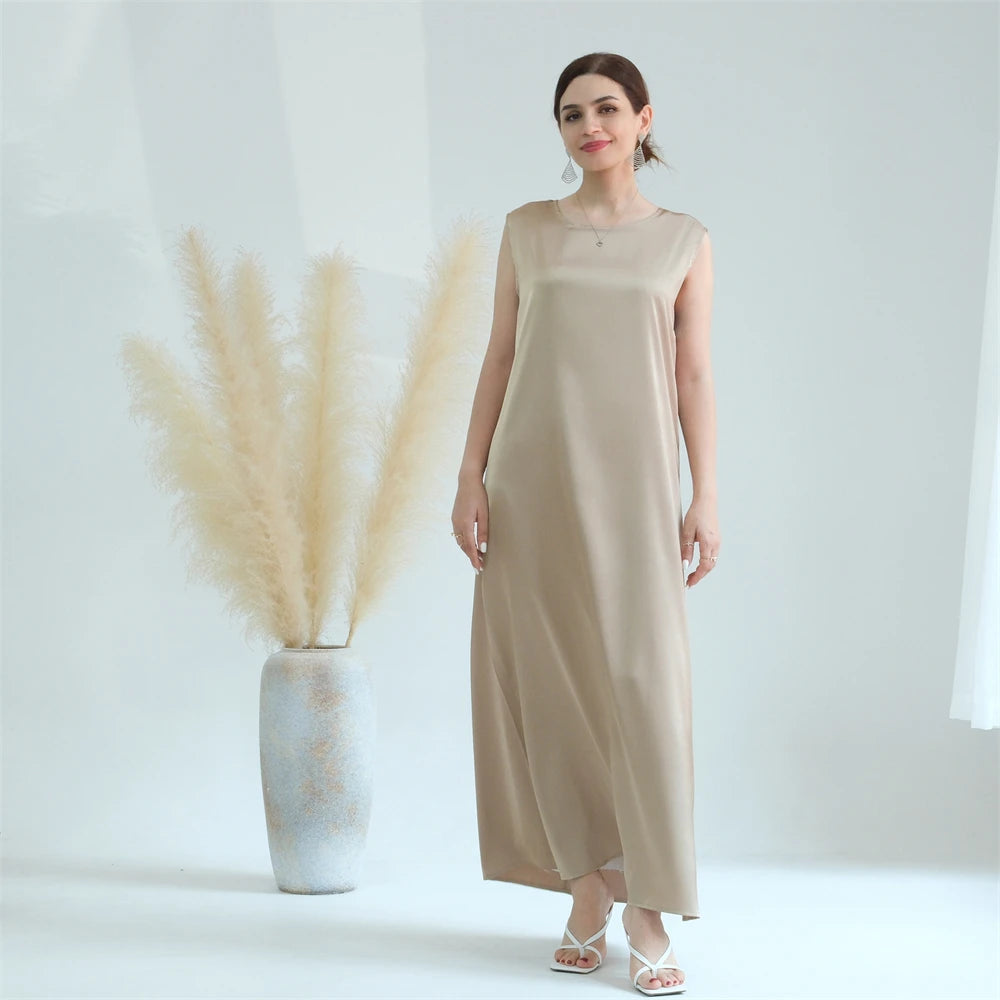A woman wearing a beige flowing modest abaya dress stands next to a vase of dried pampas grass.